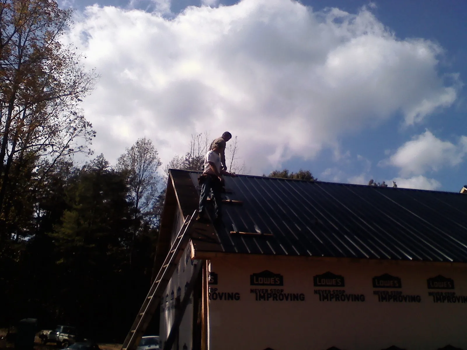 Roofer fastening dark metal panels high against a cloudy sky, from a Roof Inspection job in Shirley